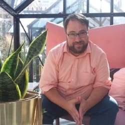 A photograph of Jer Staes in a pale pink shirt next to a green plant within an outdoor glass enclosure in the background.