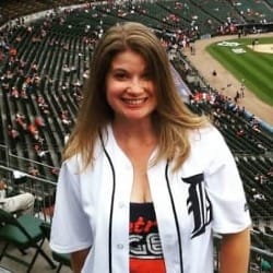 A photograph of Robin Runyan looking into the camera wearing a white Detroit Tigers jersey with the Comerica Park stands in the background.